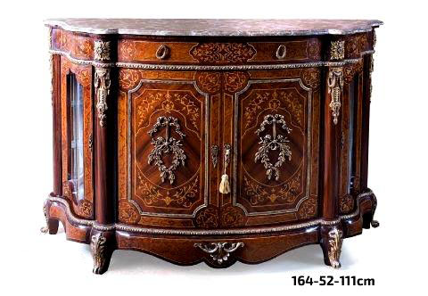 An opulent French Transitional style ormolu-mounted marquetry Sideboard, The eared beveled serpentine shaped marble top above a conforming frieze marquetry and veneer inlaid with central large drawer has two oval ring ormolu handles, the serpentine curved sides separated by circular supports ornamented with large ormolu rosettes above a chiseled brass trim, The central part with two central doors sans traverse veneer and marquetry inlaid with floral and scroll patterns, decorated with exquisite and finely chased ormolu mounts of knotted ribbons hanging a swaging foliate laurel wreaths, all within a dentil ormolu band, The circular supports with extremely finely chiseled ormolu royal chutes and two foliate ormolu keyhole escutcheons, The sides with two serpentine shaped veneer and marquetry doors with floral ormolu keyhole escutcheons, the two doors open to reveal two glass shelves, The lower serpentine scalloped veneer inlaid apron ornamented with Egg and Dart style ormolu trim, central foliate scrolled acanthus ormolu mount, the short cabriole feet adorned with scrolled acanthus ormolu sabots