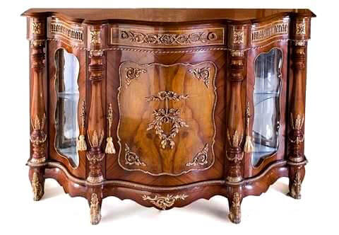 A robust French Napoléon III style distressed ormolu mounted sans-traverse veneer inlaid serpentine shaped display cabinet, The veneered moulded top above a frieze with central drawer ormolu decorated with Rinceau Guilloche design and two lyre shaped handles, on each side a faux drawer ornate with Lead-and-dart ormolu design; the section has four circular block adorned with large ormolu rosettes, and each is above long Baluster with repeated Astragals on top and bottom ormolu-mounted with leaves on the belly and swaging garlands on the Torus; the Abacus and the plinth ormolu surrounded as well, Between the balusters to each side a display ormolu bordered glass door;  The central balusters flanking a central sans traverse veneer inlaid door ornamented with beaded ormolu frame surrounding corner acanthus volutes around a central intricate and finely chiseled ormolu mount of knotted ribbons hanging a swaging foliate imbricated Laurus Nobilis, The lower arbalest shape frieze is topped with an ormolu Leaf-and-Dart trim, centered with ormolu pierced C scrolled acanthus volute and raised on four short cabriole legs ornamented with acanthus ormolu sabots