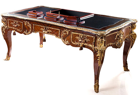 Fine French Regency Louis XV style gilt-ormolu-mounted veneer inlaid three drawers Bureau Plat after the model by Leon Kahn after the model designed by Charles Cressent, Late 19th century, the rectangular gilt-tooled red leather-inset top within a molded surround with floral and rocaille-cast clasps to each corner, over a frieze with three drawers to the front and three false drawers to the reverse, sides ornamented with ormolu frames and centered by an Apollo mask, each corner is mounted with a finely chiseled mask, on espagnolette-headed splayed legs terminating with acanthus foliate sabots, with a matching chair available per request
