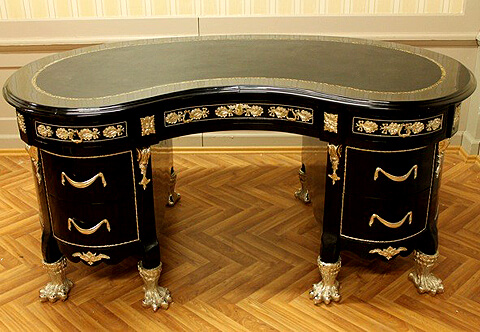 A unique Louis XVI style kidney-shaped gilt-ormolu-mounted black colored desk on the manner of Guillaume Benneman craftsmanship, Late 19th century, The beveled fitted gilt-tooled leather top above the apron contains three drawers, the central concave long drawer with ormolu foliate keyhole escutcheon, flanked by two convex drawers with lyre shaped ormolu handles, all drawers ornamented with ormolu knotted-ribbon oak-leaves and separated by blocks decorated with ormolu rosettes, The above apron surmounting four deep drawers, two to each side, bordered with a hammered ormolu band and drape shaped ormolu handles terminating with scalloped shaped frieze ornate with foliate design ormolu mount, and headed with gilt-ormolu acanthus volute bracket shaped chutes to each corner, The desk is raised on astonishing massive ormolu acanthus-sheathed hairy paw feet, The back and curved sides are plain bordered with hammered ormolu band and centered with fine ormolu capuchon