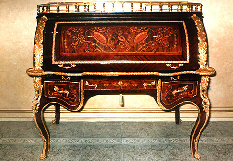 French Louis XV period style marquetry, veneer inlaid and ormolu-mounted Bureau à Cylindre, The bureau is raised by four cabriole legs decorated by ormolu chutes extended with foliate ormolu band to the paw feet sabots, The chutes flanking to the scalloped apron with three drawers framed with hammered ormolu borders and ormolu foliate handles, inlaid with foliate marquetry patterns, the central drawer with ormolu keyhole escutcheon, The side drawers are separated by a long acanthus gadroon shaped ormolu chutes extending with ormolu border elongated to the internal side of the cabriole legs, The frieze of drawers is topped with a gilt-tooled leather fitted pullout sliding writing surface with foliate ormolu handles, Above is the roll top cylinder, the curved sides ornamented with rich wide ormolu acanthus movements terminating with leafy ormolu clasps, all above a scalloped shaped surface adorned with a Cyma Recta style ormolu band all around the contour, The roll top door is inlaid with marquetry patterns of musical instruments and scrolled leafy ornamentations inside a fillet, sans traverse palisander veneer inlay and Cyma Recta style ormolu band, The roll top opens with two ormolu foliate handles to eight different size small drawers, one central door and two compartments, all decorated with marquetry works of foliate and musical trophies patterns, ormolu borders, ormolu cartouches with scrolled leaves.
The roll top is surmounted with a gilt-ormolu baluster shaped pierced three quarter opened gallery, The convex shaped sides inlaid with sans traverse veneer inlays and marquetry patterns of musical trophies and foliate scroll-works
