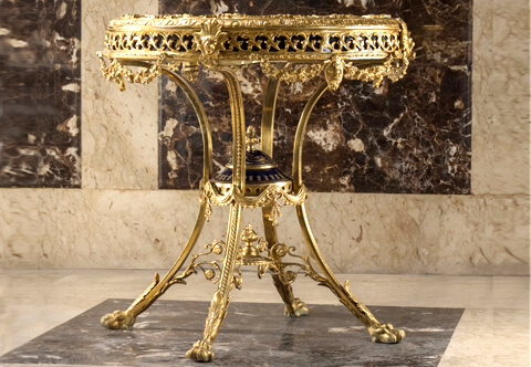 A spectacular and most impressive Napoleon III period Neo-Classical style professionally chiseled ormolu and sevres porcelain mounted Royal Center Table; the top is inset with a fine cobalt-blue ground miniature of royal court scene surrounded by fourteen porcelain miniatures of courtly members, all within an engraved gilt-ormolu surrounding border of Egg-and-Dart trim above a foliate guilloche gallery adorned with sunburst lion masks and blossoming laurel wreath pendants; raised on splendid four curved legs with acanthus leaves paw feet connected by a fine scrolled stretcher and a most decorative central reserve with an eternal fire finial; surmounted with finely chased ormolu drapes and cobalt blue porcelain central piece with lid adorned with another eternal fire finial